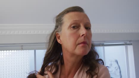 Senior-caucasian-woman-standing-in-room-looking-in-mirror-and-brushing-her-hair