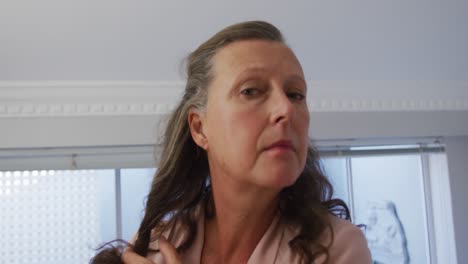 mujer caucásica mayor de pie en la habitación mirándose en el espejo y cepillándose el cabello