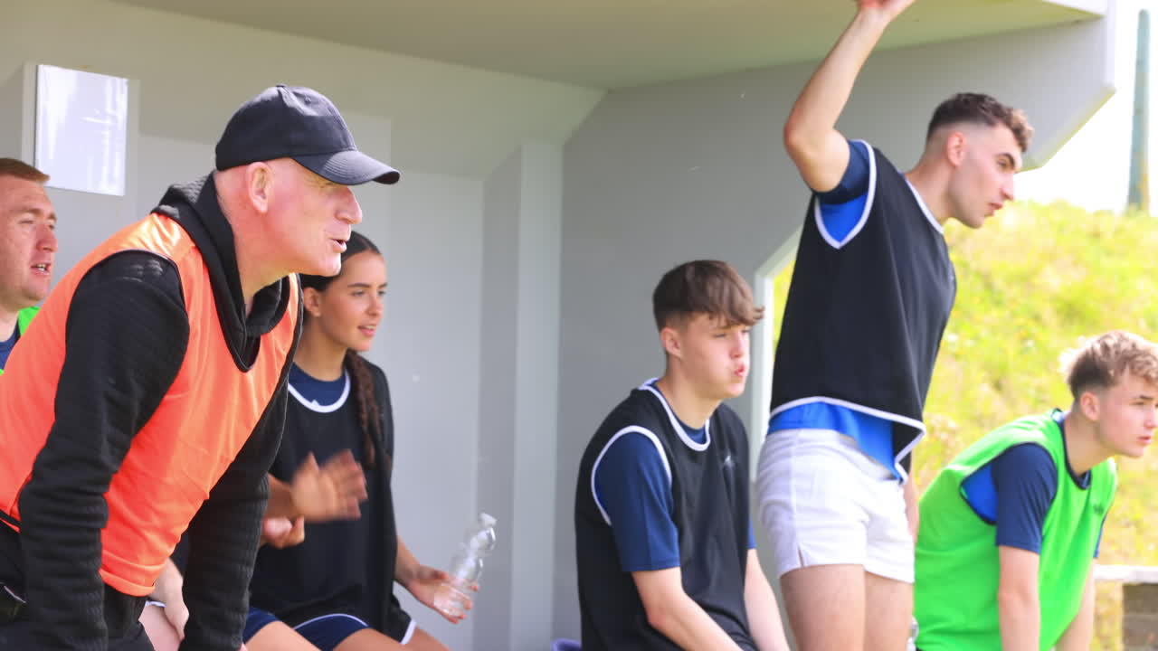 Male and female soccer players sitting on bench, shouting and supporting with coach on pitch