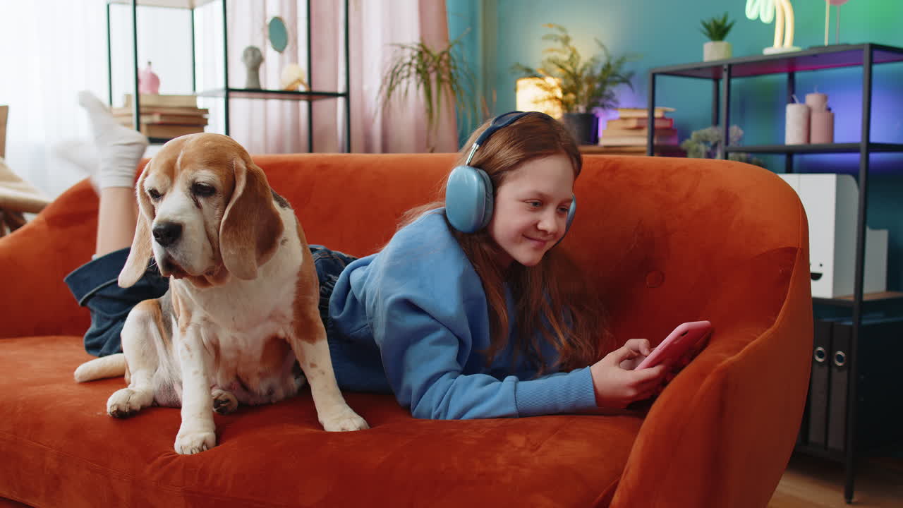 niña niño en auriculares inalámbricos relajándose usar teléfono inteligente tendido en el sofá en casa cerca de perro beagle