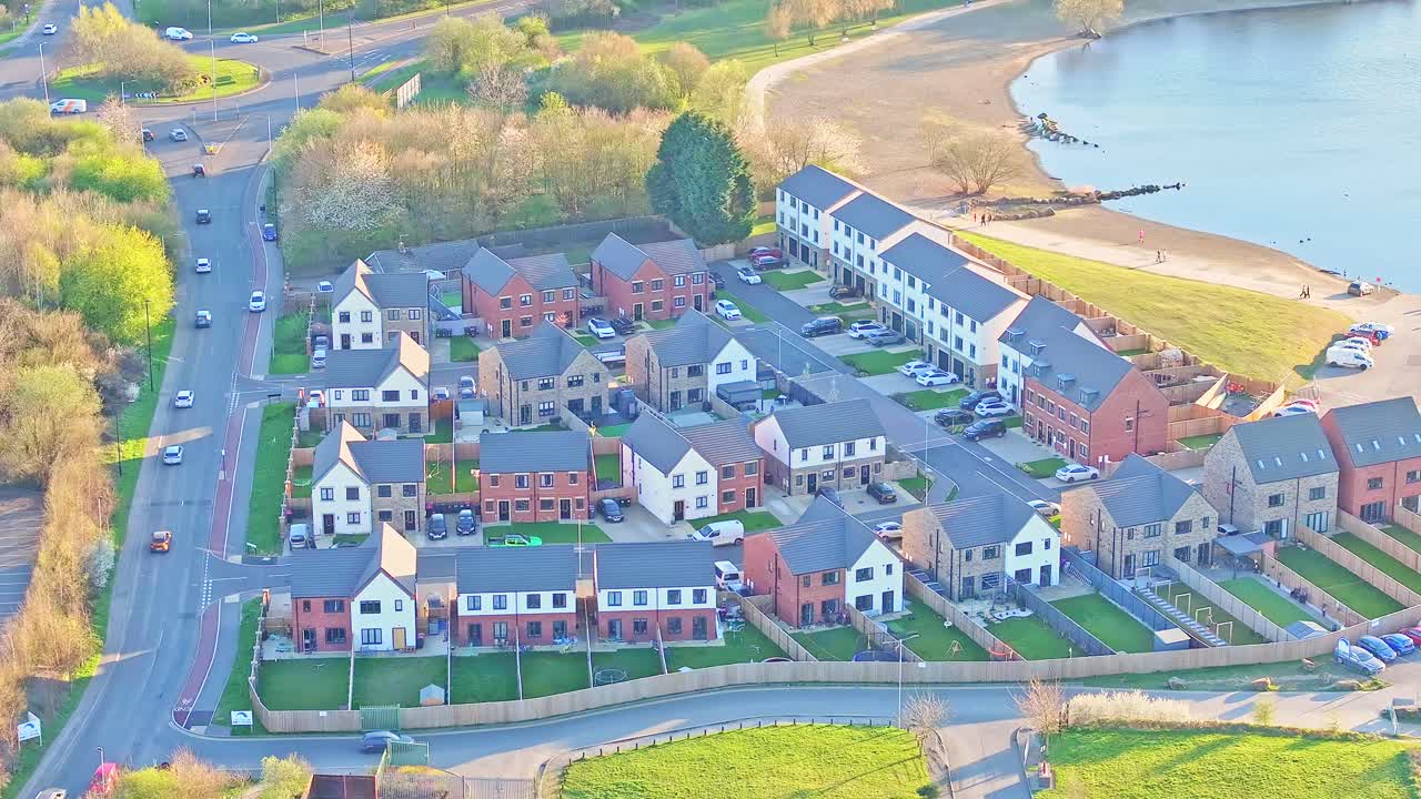 Drone view shows colorful modern houses in a quiet residential area near the beach of Manvers Lake in Rotherham, surrounded by early spring trees and morning light, real time