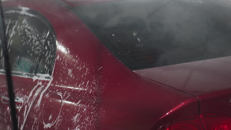 rear view of person using high pressure hose to rinse thick soap foam from red car rear inside garage environment showing suds dripping and water streams flowing dynamically over vehicle surface