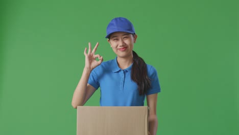 푸른 유니폼을 입은 아시아 여성 택배기사는 스튜디오에서 녹색 스크린 배경에 상자를 배달하는 동안 은 몸짓과 미소를 짓고 있습니다.
