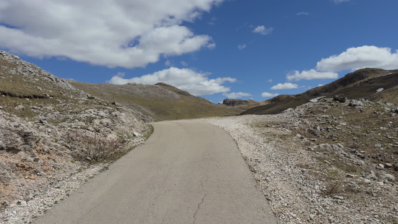 A winding, narrow mountain road curves through a dramatic landscape beneath a blue sky dotted with clouds. Captured in a wide shot and slow motion on a sunny day