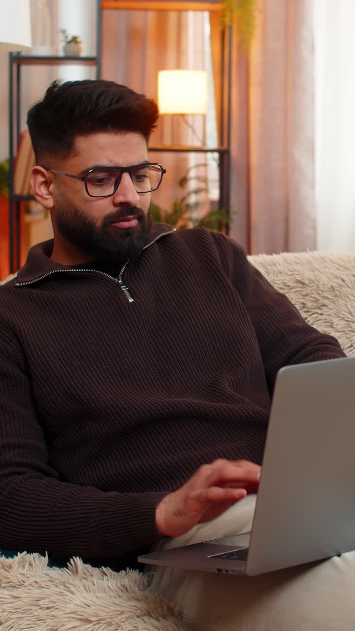 Indian sad man massaging neck on sofa with laptop at home tired from long hours of screen work
