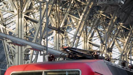 Tren-Rojo-Con-Pantógrafo-Bajo-Una-Intrincada-Estructura-De-Puente-Metálico,-Primer-Plano-Detallado-Del-Conector.