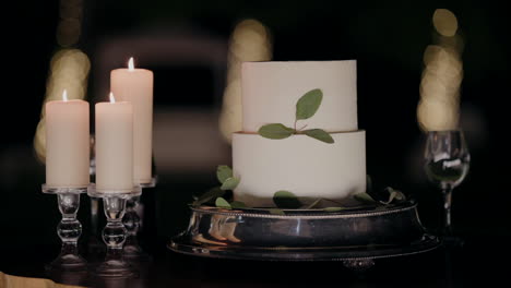 Elegante-Pastel-Blanco-De-Dos-Niveles-Adornado-Con-Hojas-Verdes,-Exhibido-En-Una-Bandeja-De-Plata-Con-Velas-Encendidas-Al-Lado,-Sobre-Un-Fondo-Ambiente-Oscuro