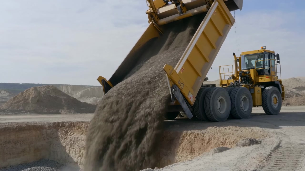 Yellow Dump Truck Unloading Gravel Into Deep Pit At Open Quarry, Heavy Truck Tilts Bed To Release Coarse