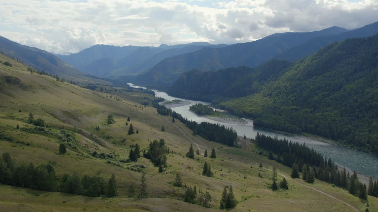 Wide aerial landscape of Ak-Kem valley with a winding river, forested slopes, and layered mountains under cloudy sky in Altai Republic, Russia