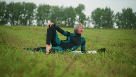 woman lying on her side with hand resting on her leg, smiling peacefully, with a background of greenery and trees in a wide, open grassy field under a cloudy sky