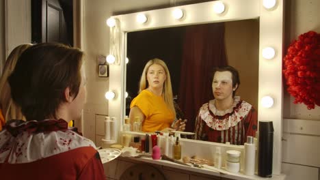 woman applies clown makeup to a man in a dressing room