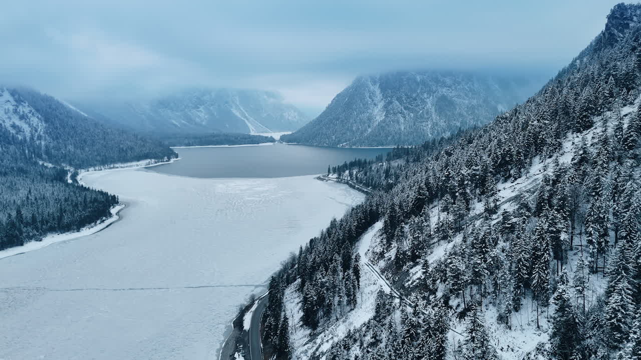 Slope of the mountain descending into the lake is covered pine tree woods. Picturesque winter rocky landscape from drone.