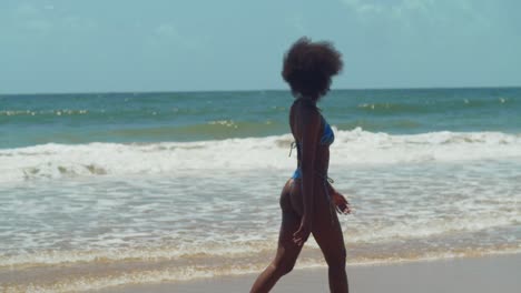 in the midst of a tropical paradise, a girl with curly hair walking on the shoreline on a caribbean island beach in a bikini with ocean waves in the background