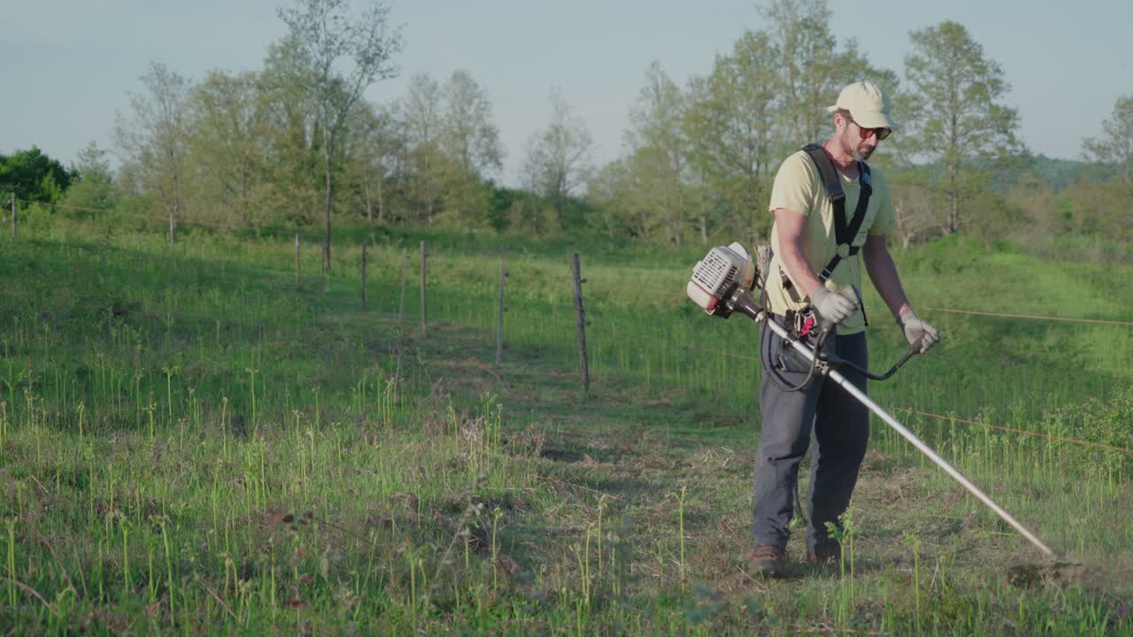 A man wearing protective gear uses a gasoline brush cutter to trim tall grass and weeds in a green rural field