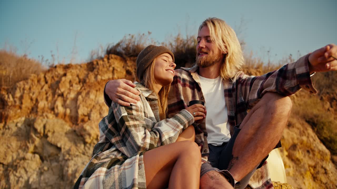 Happy couple, a blond man with a beard in a checkered shirt smokes a cigarette while sitting with his blonde girlfriend in a checkered shirt and a green hat on a large stone on a rocky shore near the sea and points somewhere towards the sunrise in the morning near their paddle boards surf