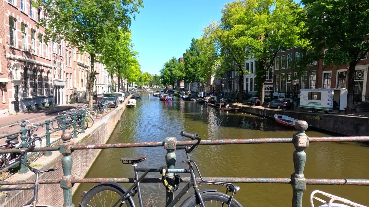 Static wide shot of Amsterdam canal, bicycles on bridge, summer daylight, vibrant cityscape, calm water