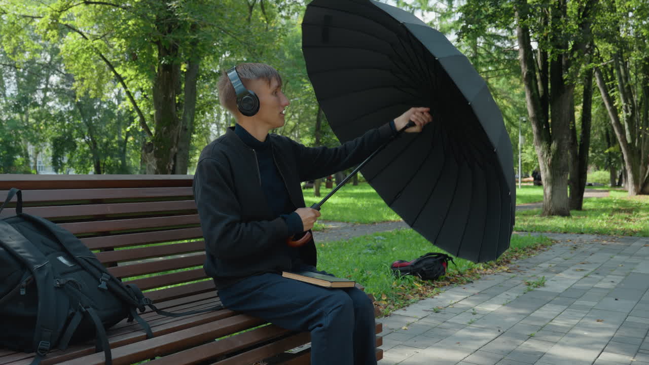 uomo su una panchina del parco che tiene un ombrello aperto, indossa cuffie, controlla lo smartphone, apre un grande ombrello nero nonostante il tempo soleggiato, zaino accanto, alberi frondosi, sentiero pavimentato, stato d'animo calmo e riflessivo, urbano