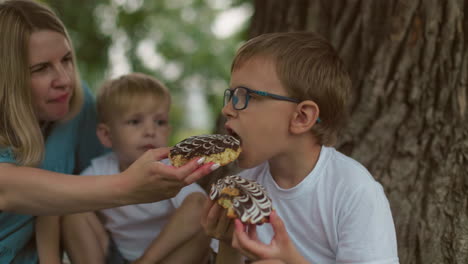 a mother shares her snack with her older son, offering him a bite while he focuses on his treat, meanwhile, the younger son watches curiously