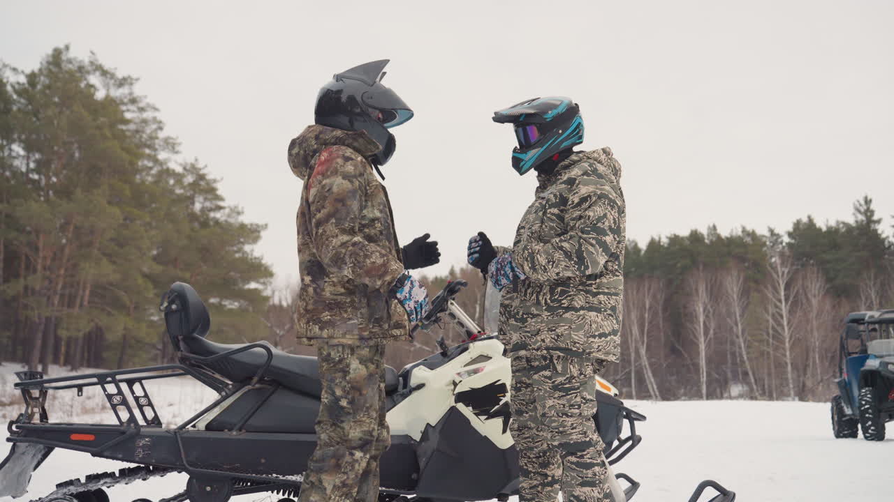 Due piloti si stringono la mano vicino alla motoslitta, tute mimetiche e occhiali a specchio, conversazione sul percorso e sui controlli di sicurezza, sagoma di un tecnico sullo sfondo vicino al confine della pineta, atmosfera di fiducia reciproca.