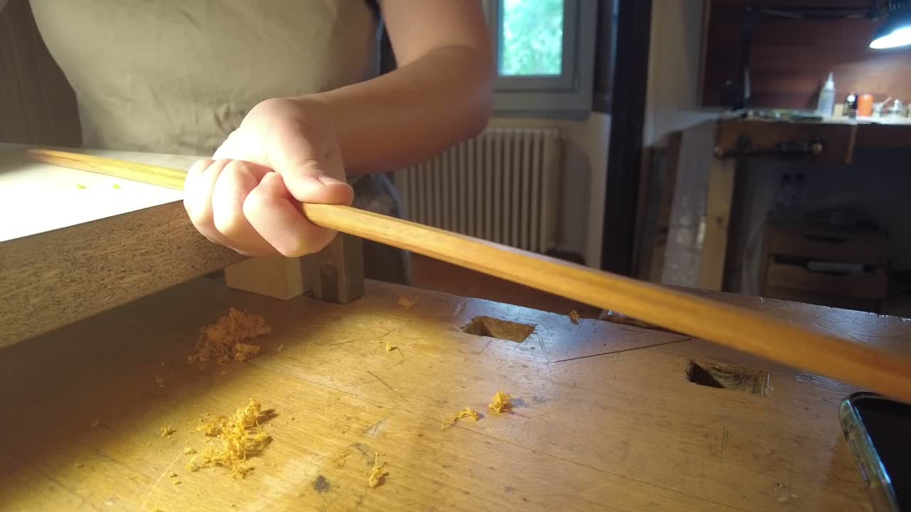 Close-up of a violin bow maker shaping a wooden bow stick on a workbench using a hand plane and wooden jig, showing traditional luthier craftsmanship and precision instrument making