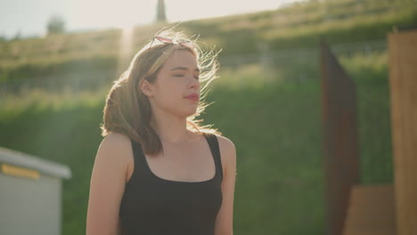 woman closes brown-covered book, takes a deep breath, and blinks her eyes while wind gently blows her hair, sunlight illuminates her face with a backdrop of greenery and wooden structures