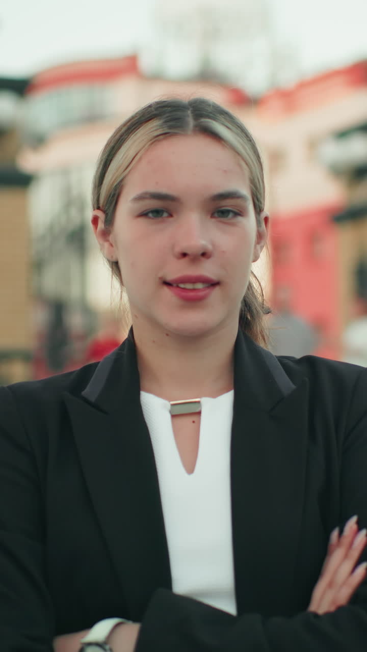 Confident woman with folded arms standing outdoors with elegant smile, dressed in business attire, exuding calm assurance and professionalism, in urban setting with blurred