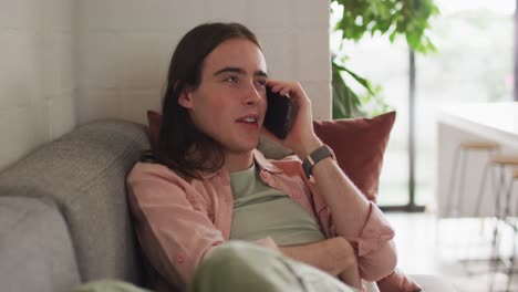 Caucasian-non-binary-transgender-woman-sitting-on-sofa,-talking-on-smartphone