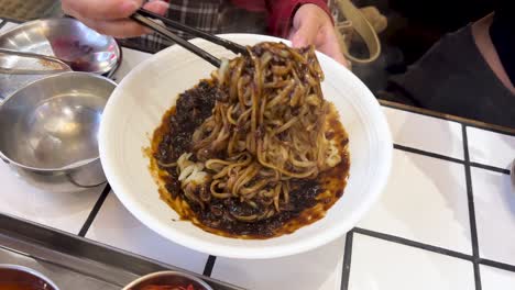 a person uses chopsticks to thoroughly mix korean black bean noodles in a white bowl, seated at a tiled restaurant table with metal bowls nearby. overhead lighting, handheld camera