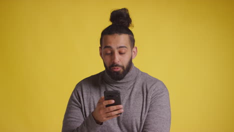 Fotografía-De-Estudio-De-Un-Joven-Sonriente-Con-Un-Teléfono-Móvil-Recibiendo-Buenas-Noticias-Sobre-Un-Fondo-Amarillo