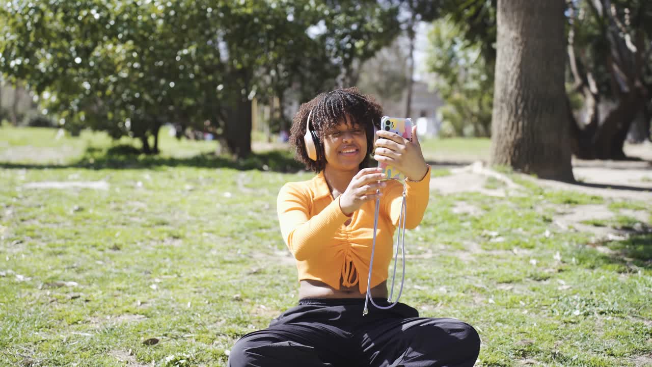 Happy black woman in headphones taking selfie on lawn