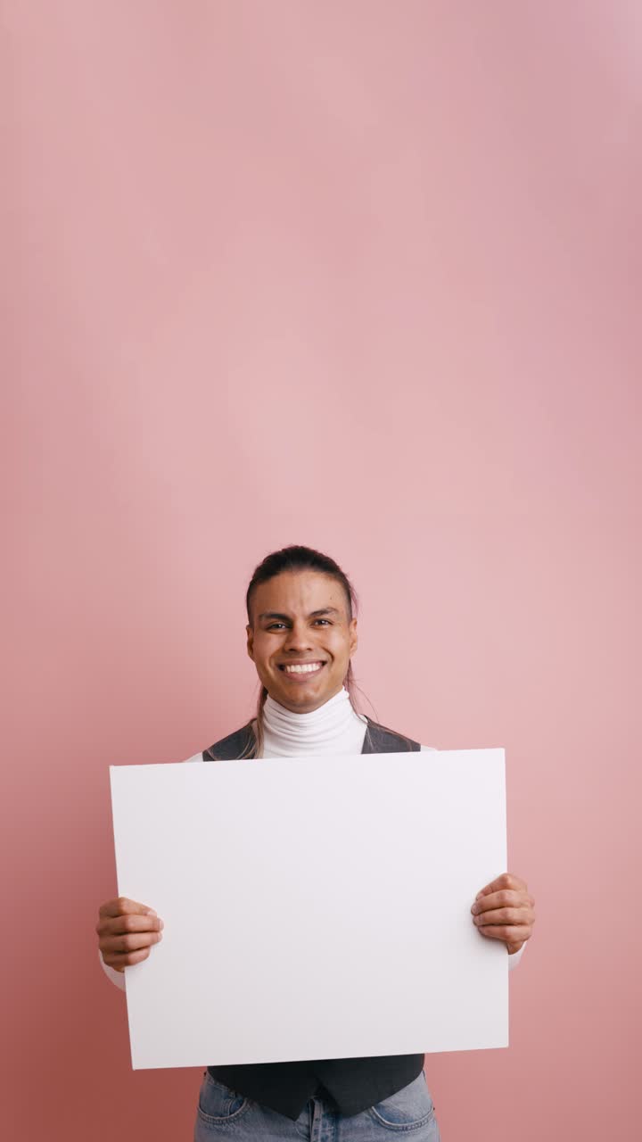 Giovane uomo sorridente che tiene in mano un cartello bianco vuoto su sfondo rosa
