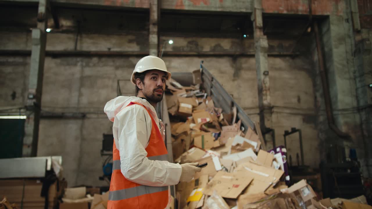 un hombre moreno con barba en un casco blanco y un uniforme blanco en un chaleco naranja se encuentra cerca de una cinta transportadora en una planta de reciclaje de papel residual