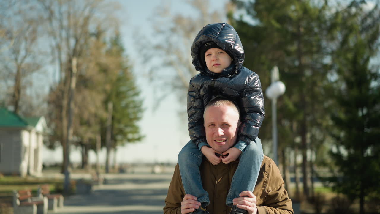 Father carrying his child on his shoulders smiling, the father is wearing a brown jacket and beanie, and the child is in a black jacket with trees and light poles behind