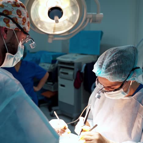 surgeon and his assistant performing cosmetic surgery in hospital operating room. surgeon in mask wearing loupes during medical procedure.