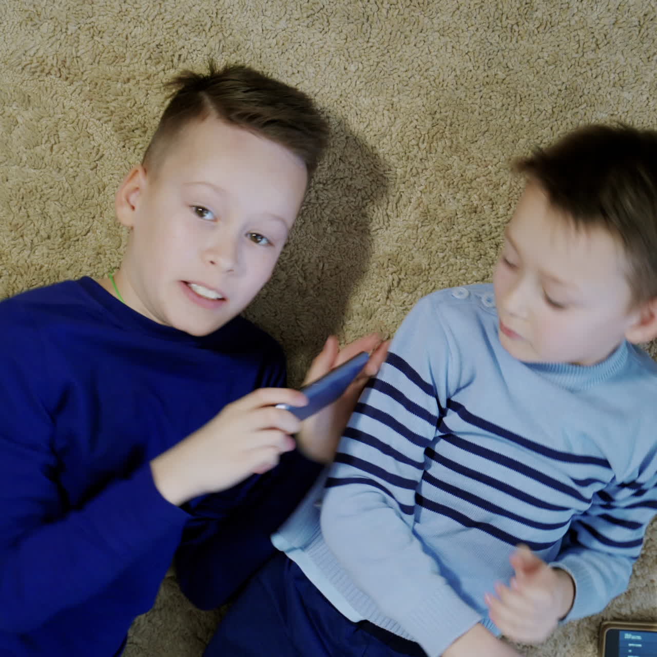 Two happy boys lying on the floor and playing on one smartphone together. Relaxed kids having fun altogether with modern gadget on a soft rug. Close-up