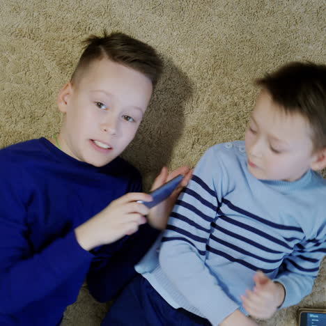 two happy boys lying on the floor and playing on one smartphone together. relaxed kids having fun altogether with modern gadget on a soft rug. close-up
