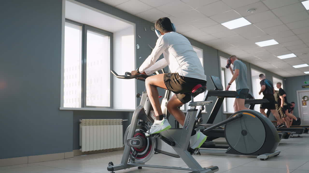 Young student in white sportswear cycles on stationary bike under bright gym lights and sunlight, while others run on treadmills and one man wipes sweat from his face with towel near window