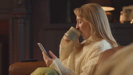 Mujer-En-Casa-En-El-Salón-Con-Un-Acogedor-Fuego-Y-Una-Bebida-Caliente-Usando-El-Teléfono-Móvil