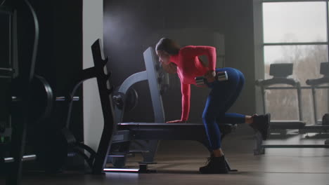 La-Mujer-Con-Un-Top-Rosa-Corto-Está-Trabajando-Boca-Arriba-En-El-Gimnasio.-Se-Apoya-En-Un-Banco-Con-La-Parte-Derecha-Del-Cuerpo-Y-Entrena-La-Mano-Izquierda-Y-La-Parte-Izquierda-De-La-Espalda.