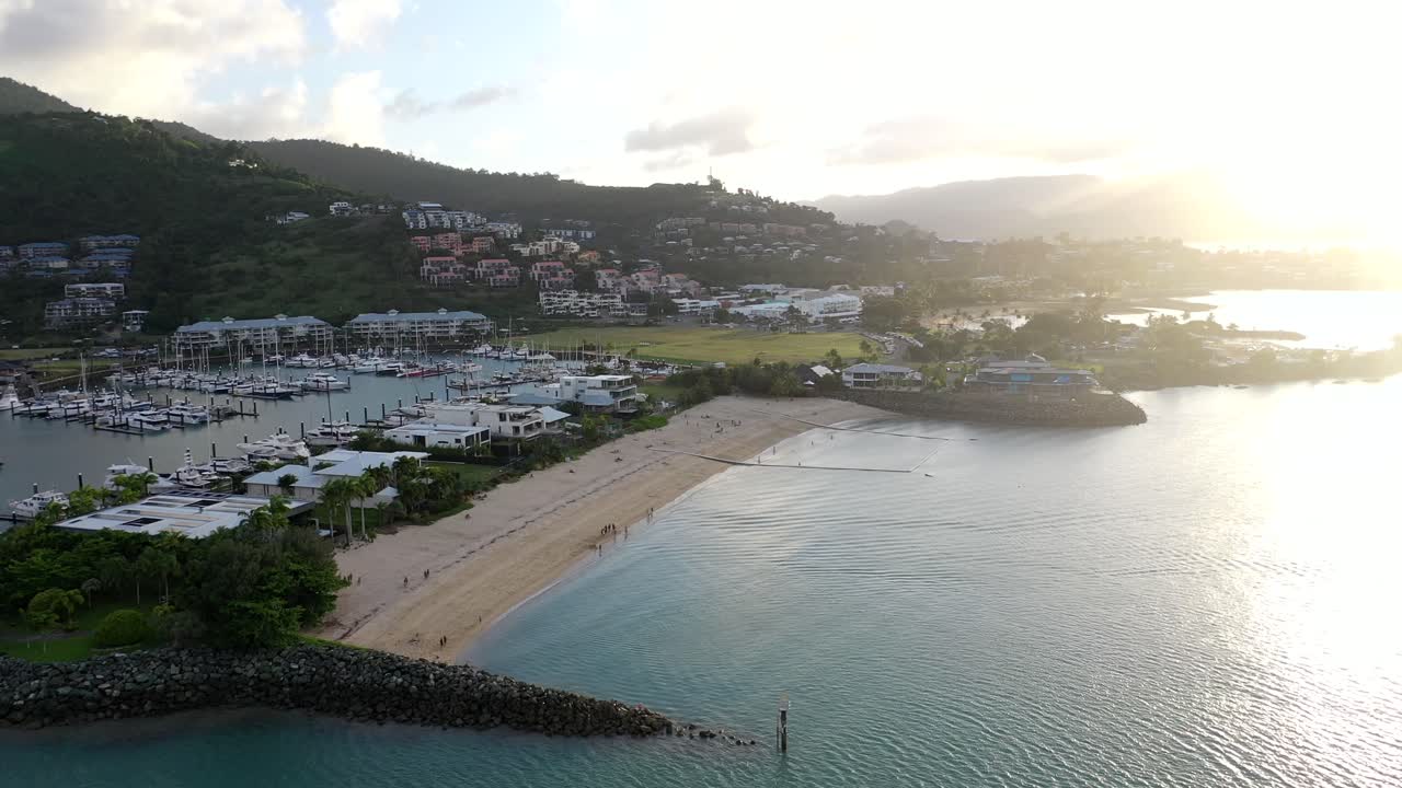 Slow pan drone shot over Airlie Beach, Queensland - showing marina area and beach, with moored yachts and boats as evening sun shines through.