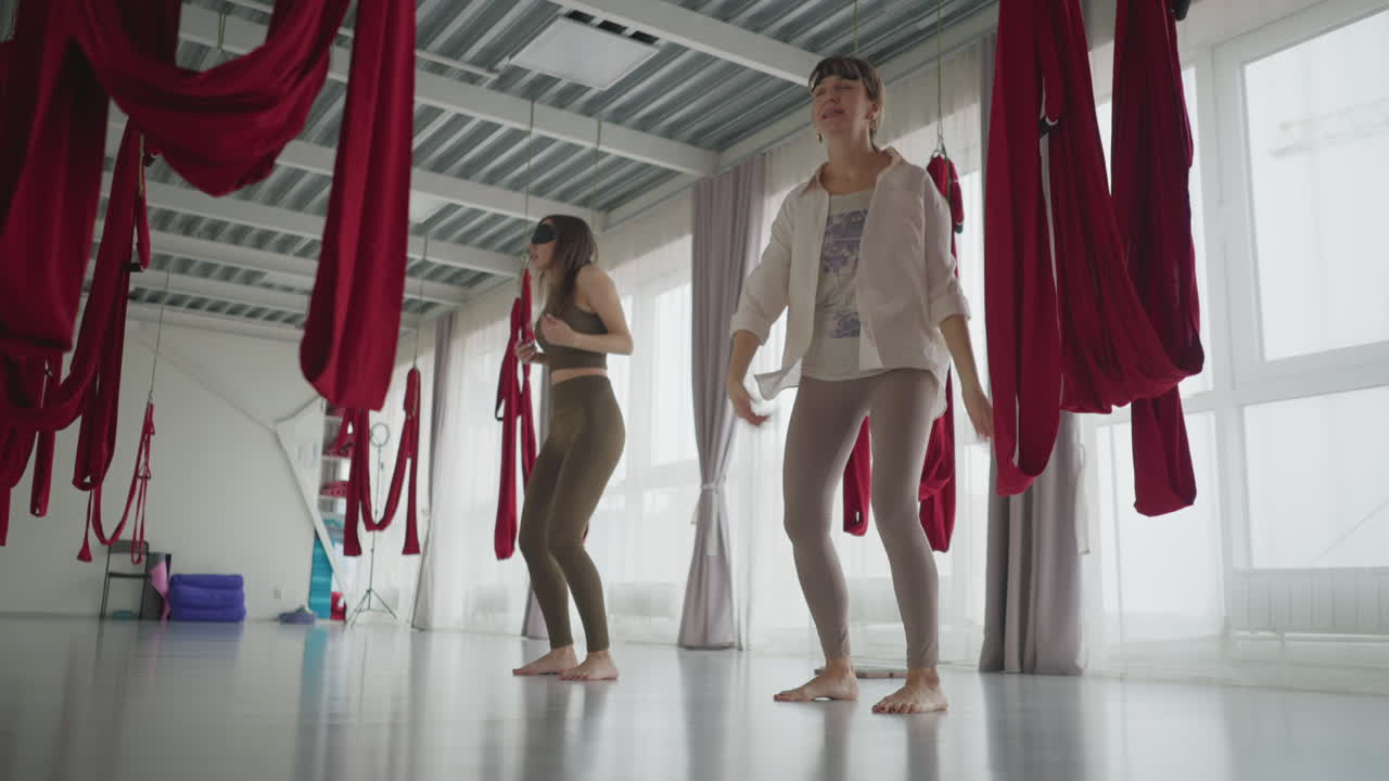 Instructors with eyes closed under hammocks perform gentle body stretch in spacious bright studio, focusing on breath and movement to release tension and enhance mindfulness through guided practice