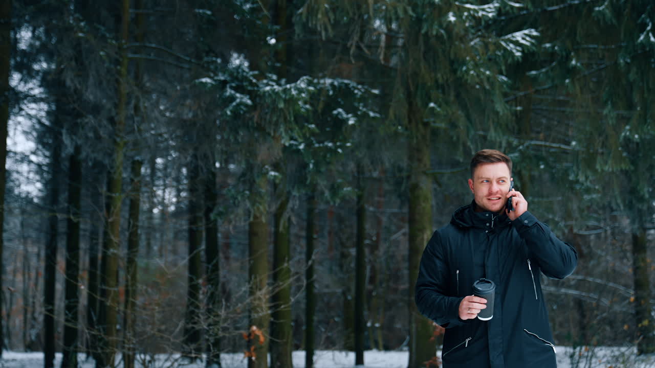 Man stands in the park holding a phone at his ear. Caucasian male with coffee cup in hand walks outdoors in winter.