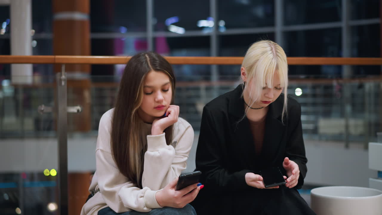 Two friends seated indoors operating phone with tired expressions in modern mall environment showing candid lifestyle mood casual fashion and natural presence
