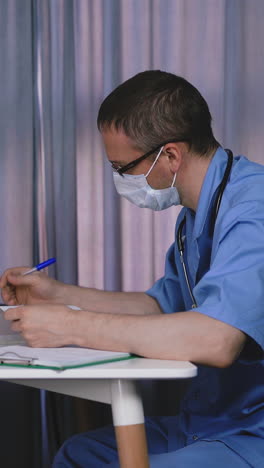 concentrated medical doctor wearing blue uniform and facial mask examines funny blonde little girl and makes notes sitting at table in hospital office