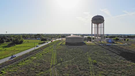 Rising-up-from-a-field-that-has-strips-of-green-grass,-flying-towards-large-water-tower-and-tank