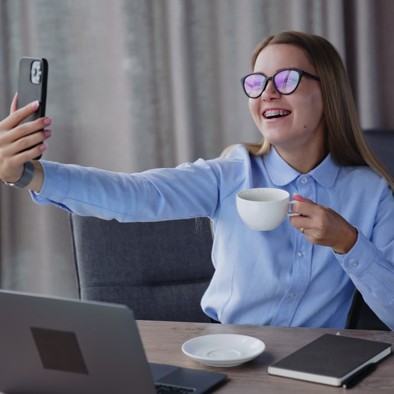 Giovane donna bionda sorridente beve un caffè seduta a un tavolo. Donna che si fa un selfie con una tazza sul telefono