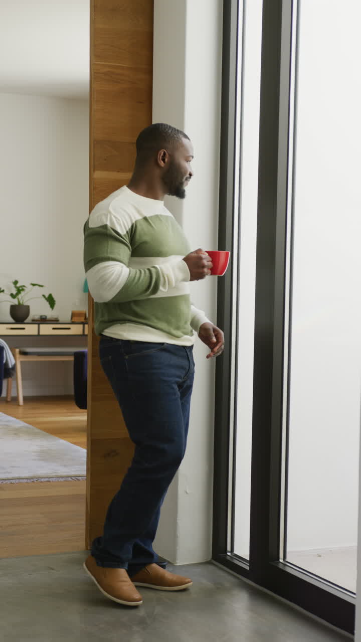 video vertical de un feliz hombre afroamericano bebiendo café y mirando por la ventana en casa