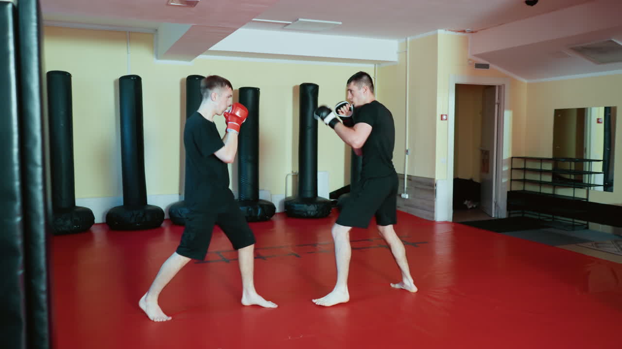 Wrestlers sparring during martial training inside gym, wearing gloves and black outfits, performing combat sport techniques with focus and movement on red mat floor surrounded by punching bags