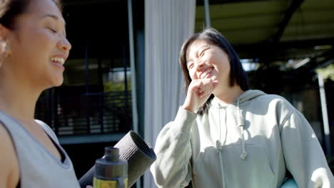 happy asian female friends with exercise mat and water bottle laughing on terrace, slow motion