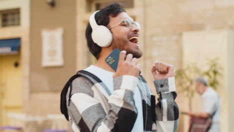 happy indian man wearing headphones choosing listening disco rock n roll music dancing on street